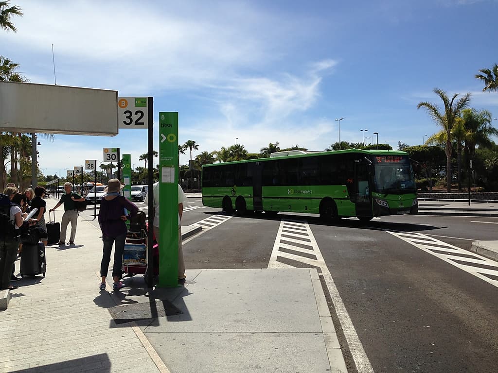 Green TITSA bus beside airport bus signs and passengers with bags.