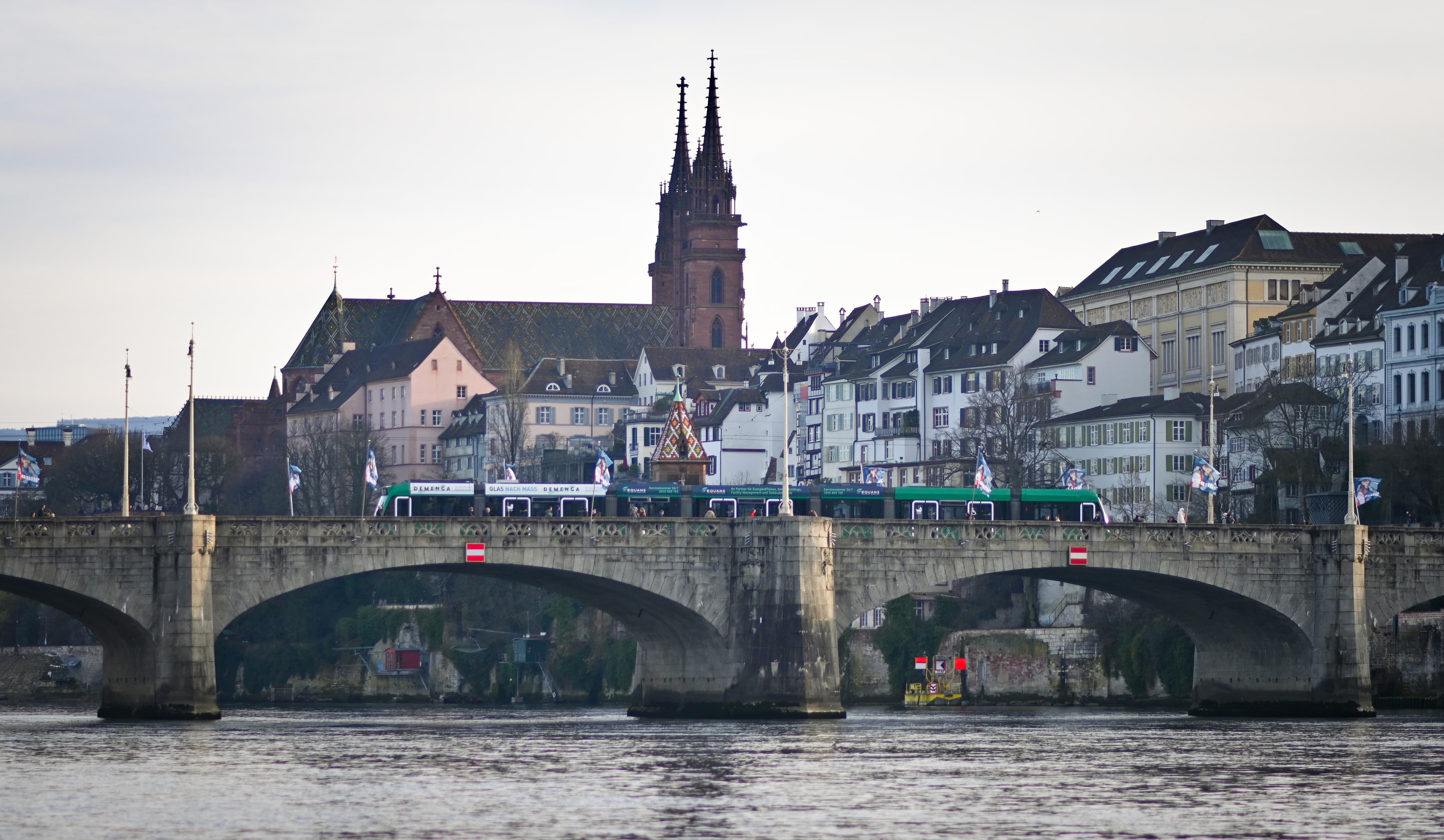 A green tram crossing Mittlere Brücke over the Rhine, with Basel Minster behind