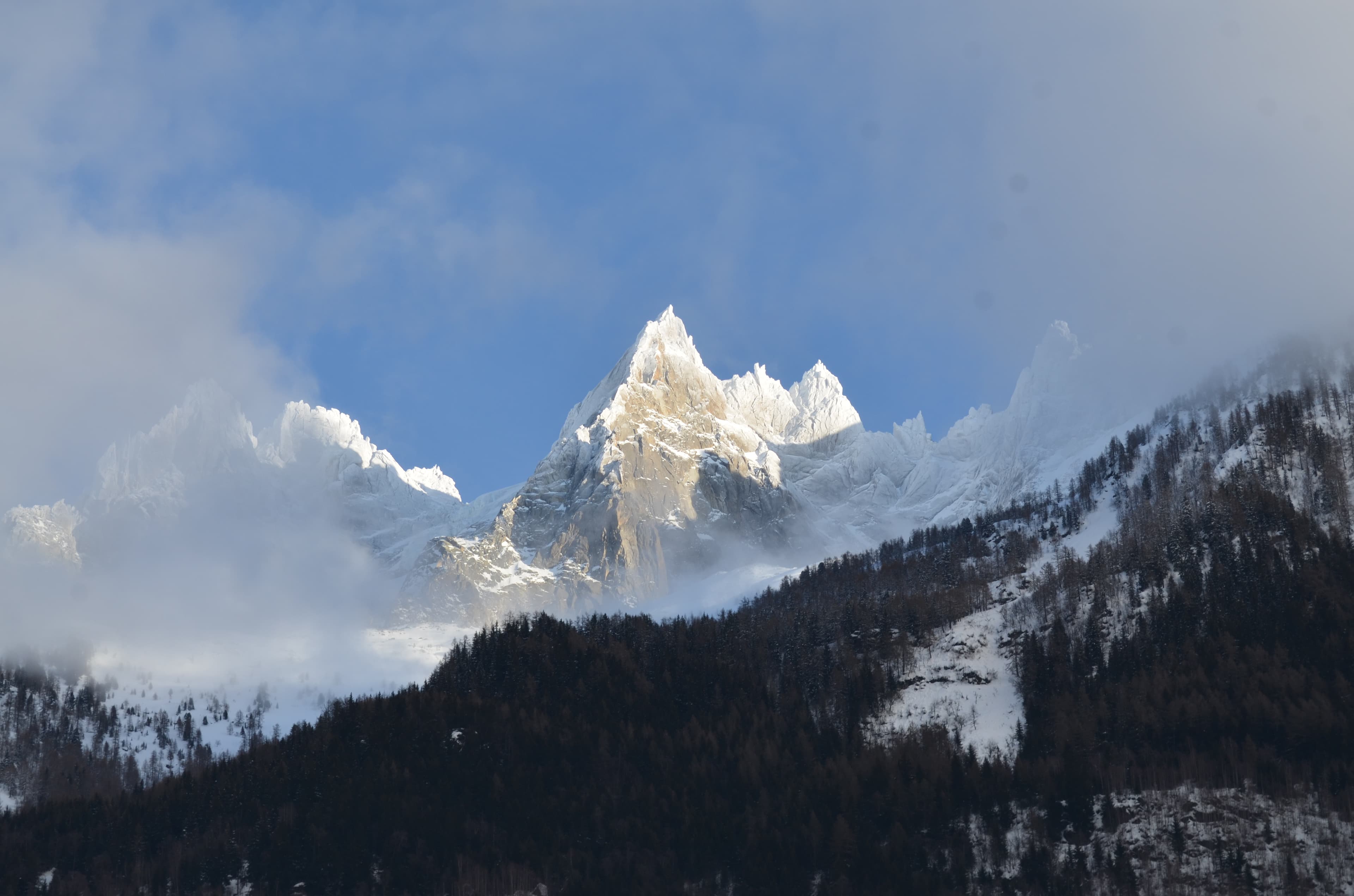 Sunlit snowy mountain slopes above Chamonix under blue sky and low cloud.