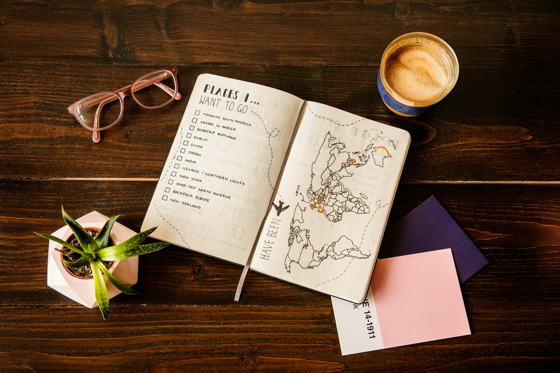 An open travel-planning notebook with a world map and checklist beside glasses and coffee.