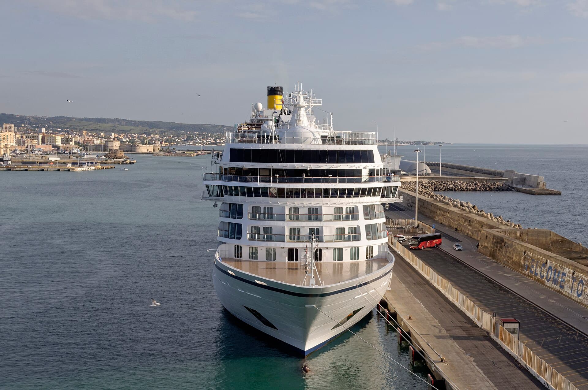 A large cruise ship moored beside a quay at Civitavecchia, with the harbour and city beyond.